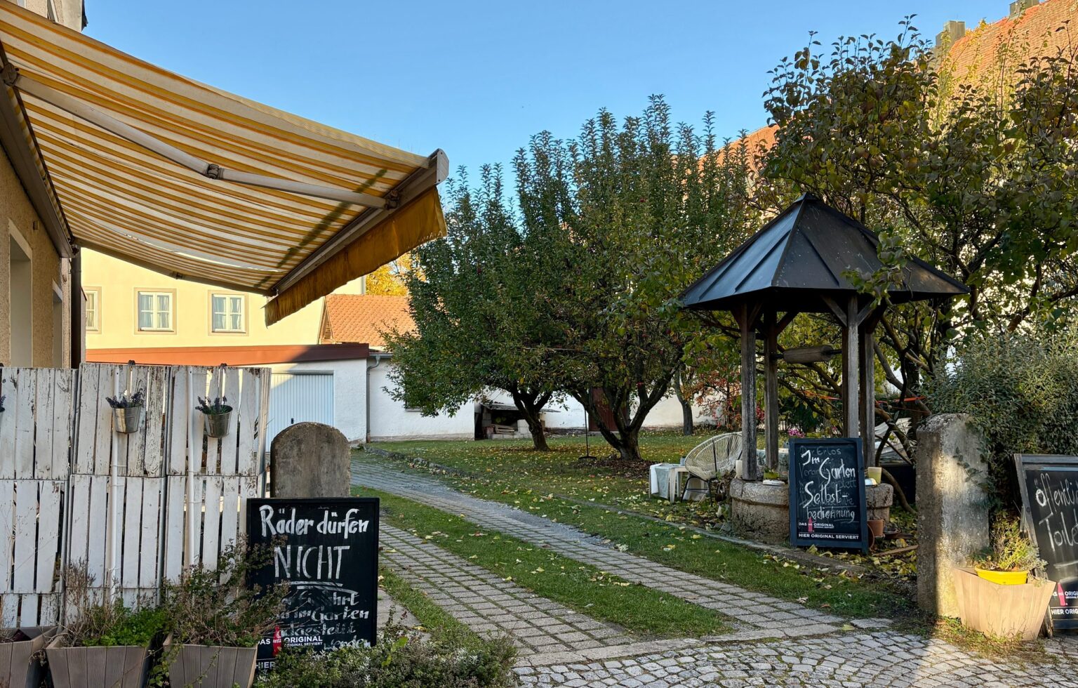 Gepflasterter Weg in den Garten des Café am Kirchplatz mit überdachtem Brunnen und Kreidetafeln